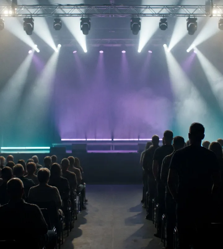An audience in silhouette facing a stage washed in violet and teal light