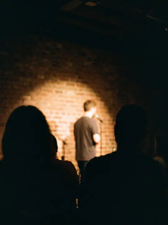 A comedian with a microphone in a single spotlight against a brick wall, the audience in silhouette