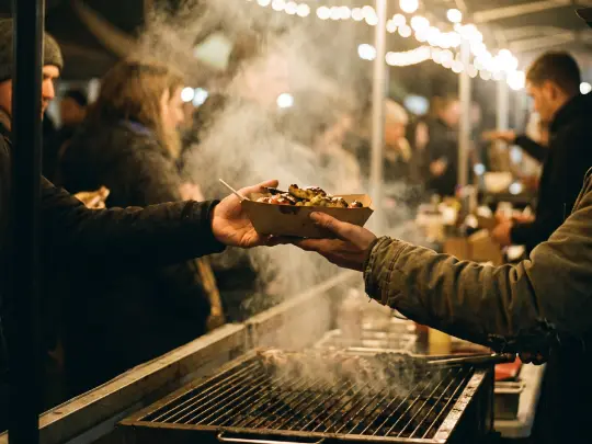 Food handed across a smoking grill at a night market under festoon lights